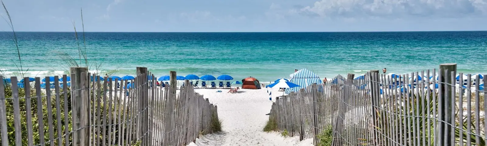 Sandy beach walkover between weathered dune fences opening to blue and white umbrellas on the Florida Panhandle coast.